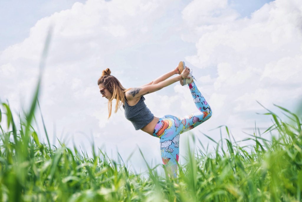 Woman with a top knot hairstyle doing a yoga pose in a grassy field against a cloudy sky