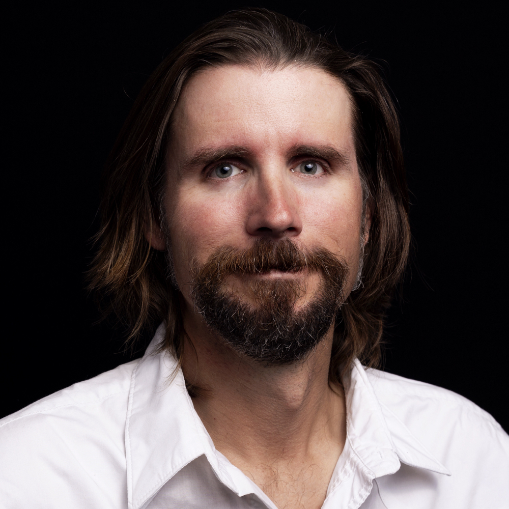 Headshot of Chris Badgett with shoulder-length brown hair and a goatee, wearing a white collared shirt